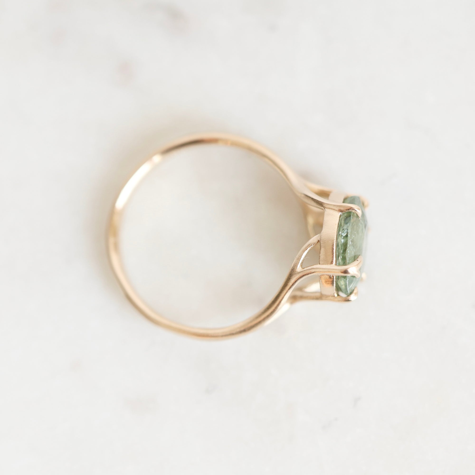 A delicate gold ring with a small green gemstone, photographed from above on a light marble surface.