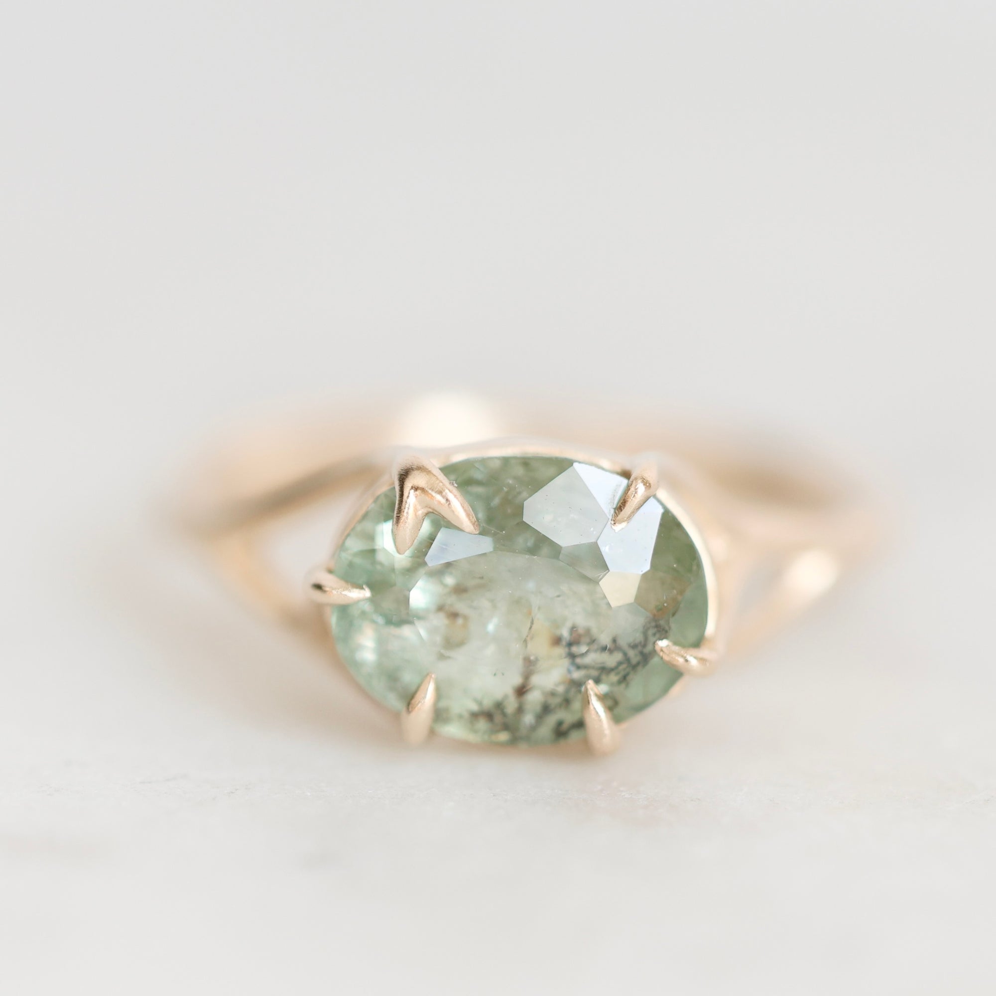 A gold ring featuring an oval-cut light green gemstone with natural inclusions, set in a four-prong claw setting on a soft, light gray background.
