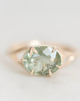 A gold ring featuring an oval-cut light green gemstone with natural inclusions, set in a four-prong claw setting on a soft, light gray background.