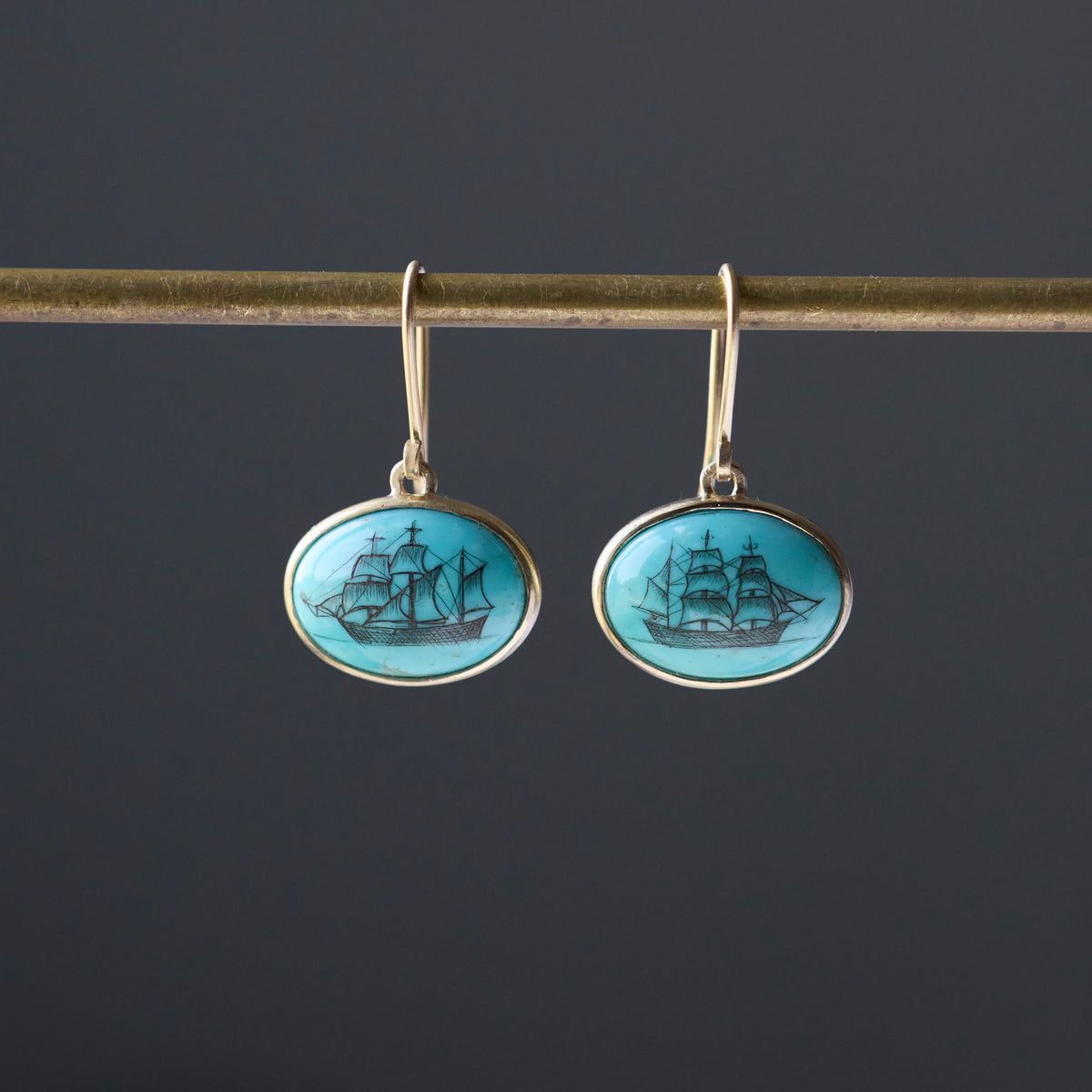 Turquoise earrings with engraved ships in black ink hang from a gold bar against a dark background. 