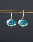 Turquoise earrings with engraved ships in black ink hang from a gold bar against a dark background. 