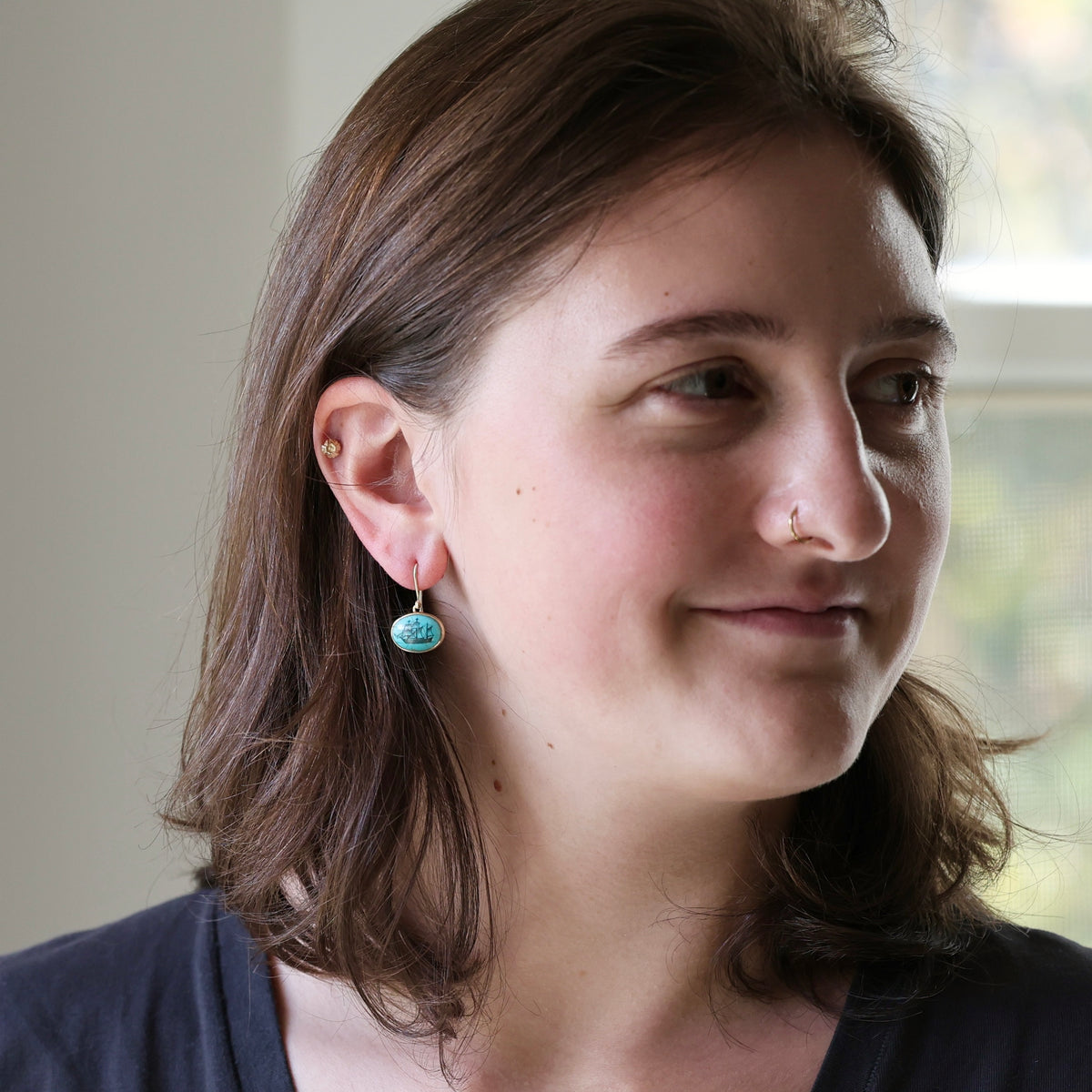 Woman wearing turquoise earring with ship engraving against a blurred indoor background. 