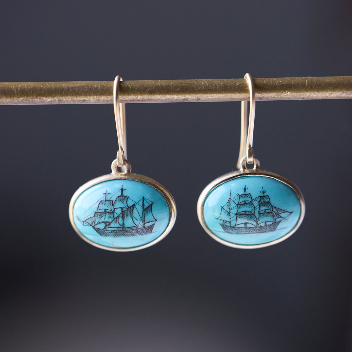 Turquoise earrings with engraved ships in black ink hang from a gold bar against a dark background. 