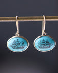 Turquoise earrings with engraved ships in black ink hang from a gold bar against a dark background. 