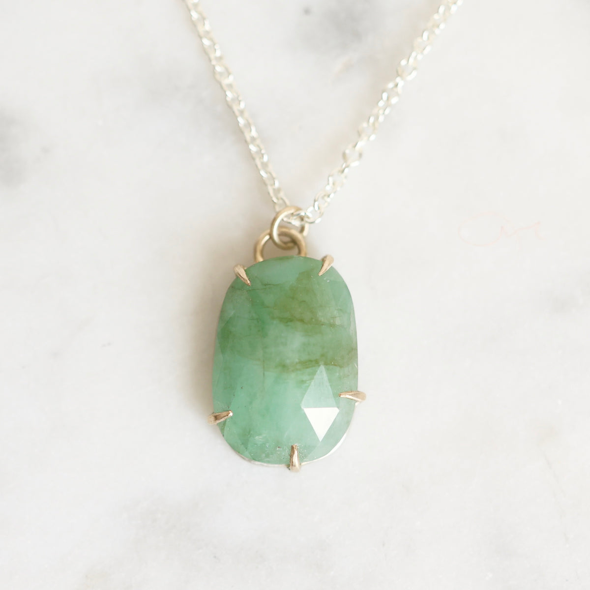 A green marquise-cut gemstone pendant in a gold bezel setting on a delicate silver chain, displayed on a light marble surface.
