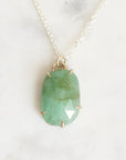 A green marquise-cut gemstone pendant in a gold bezel setting on a delicate silver chain, displayed on a light marble surface.