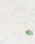 A delicate gold chain necklace with a green gemstone pendant, resting on a light gray marble surface.