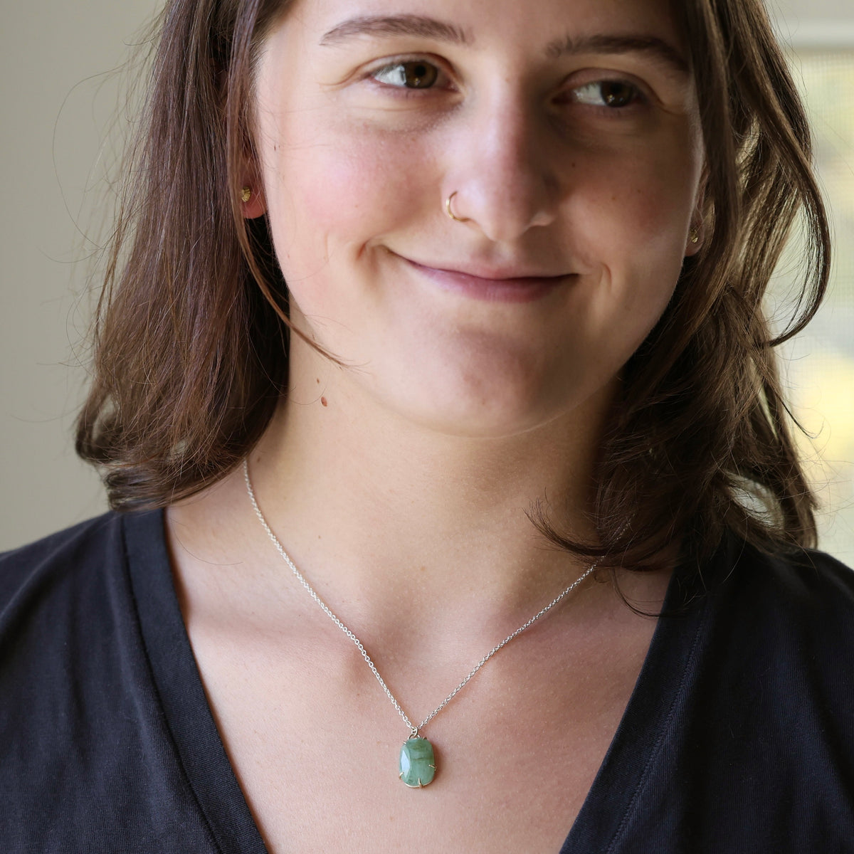 A woman with brown hair wearing a delicate silver chain necklace with a small, polished green pendant, shown in a close-up portrait.