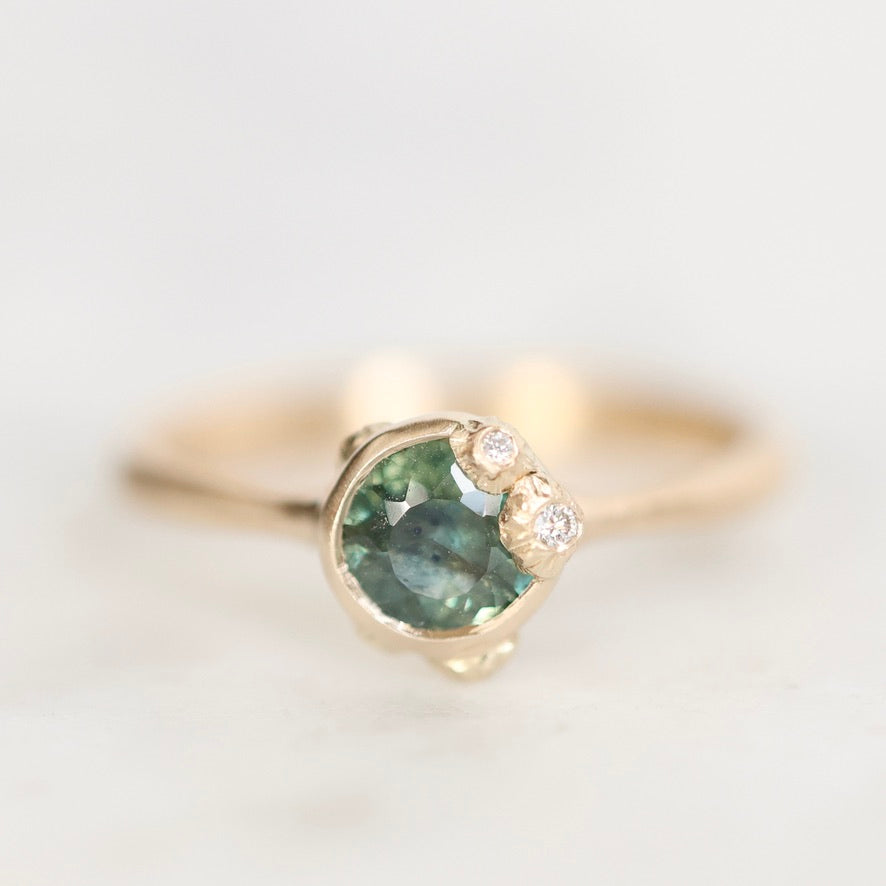 A gold ring featuring a round green gemstone in a prong setting, accented by two small clear stones on the right side, photographed on a light neutral background.