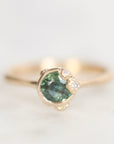 A gold ring featuring a round green gemstone in a prong setting, accented by two small clear stones on the right side, photographed on a light neutral background.