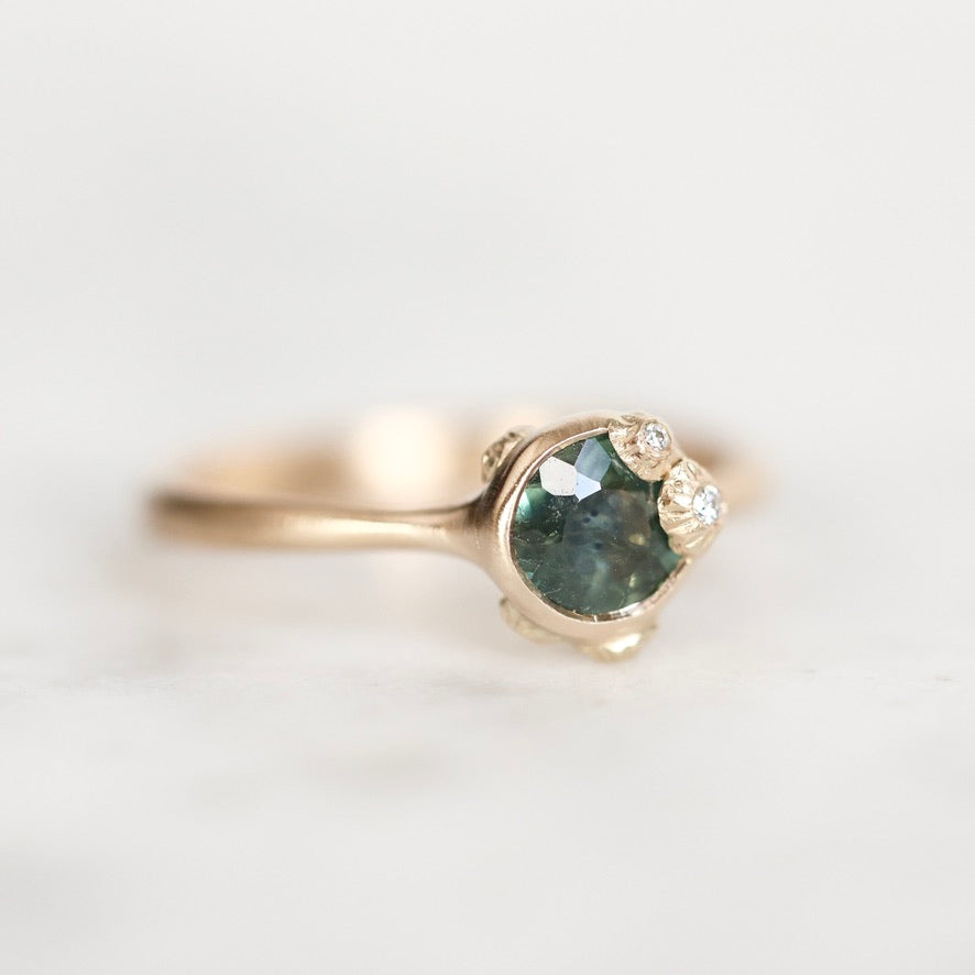 A gold ring featuring a round green gemstone in a prong setting, accented by two small clear stones, shown on a light neutral background.