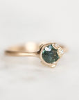 A gold ring featuring a round green gemstone in a prong setting, accented by two small clear stones, shown on a light neutral background.