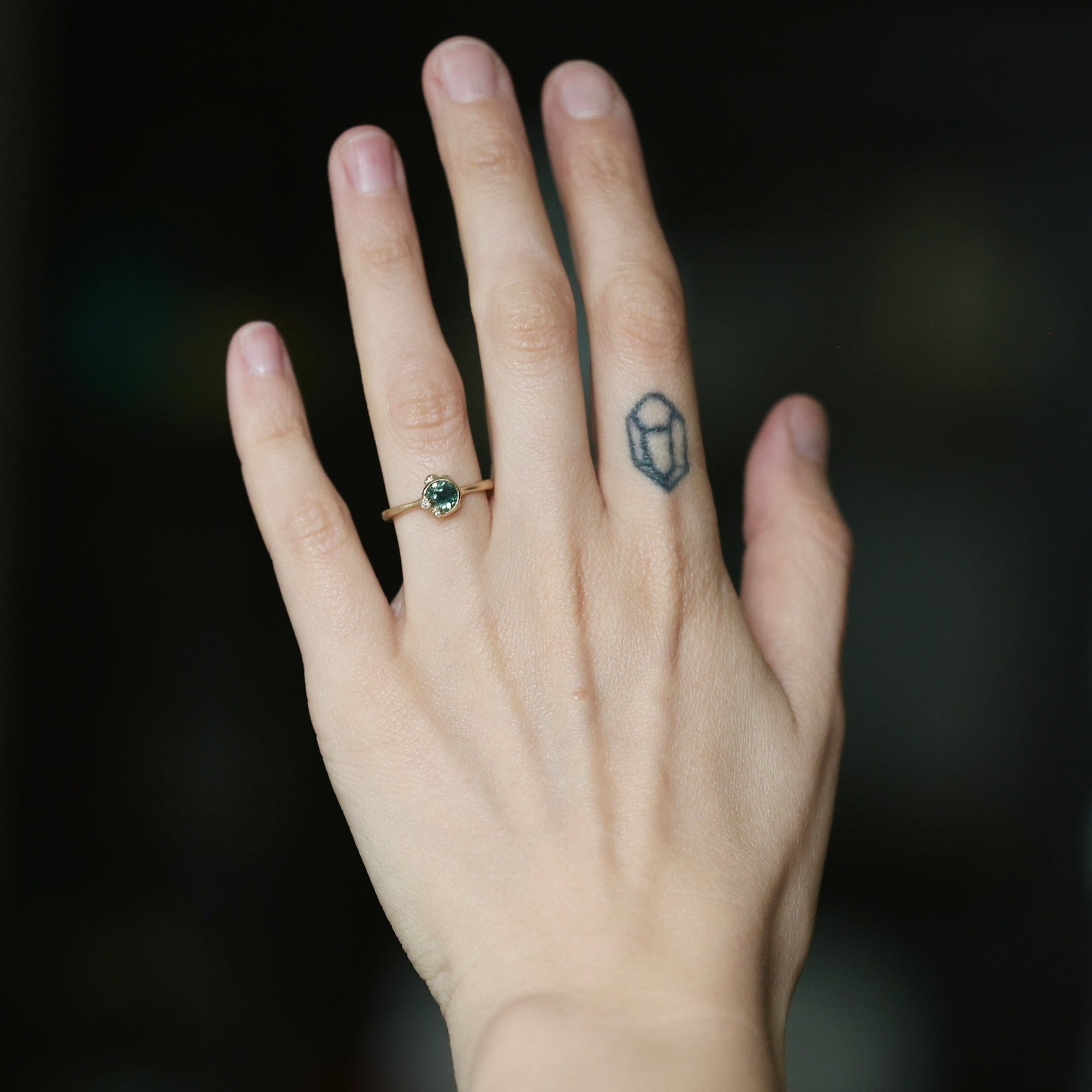 A close-up of a person's hand wearing a delicate gold ring with a small green gemstone, set against a dark, blurred background.