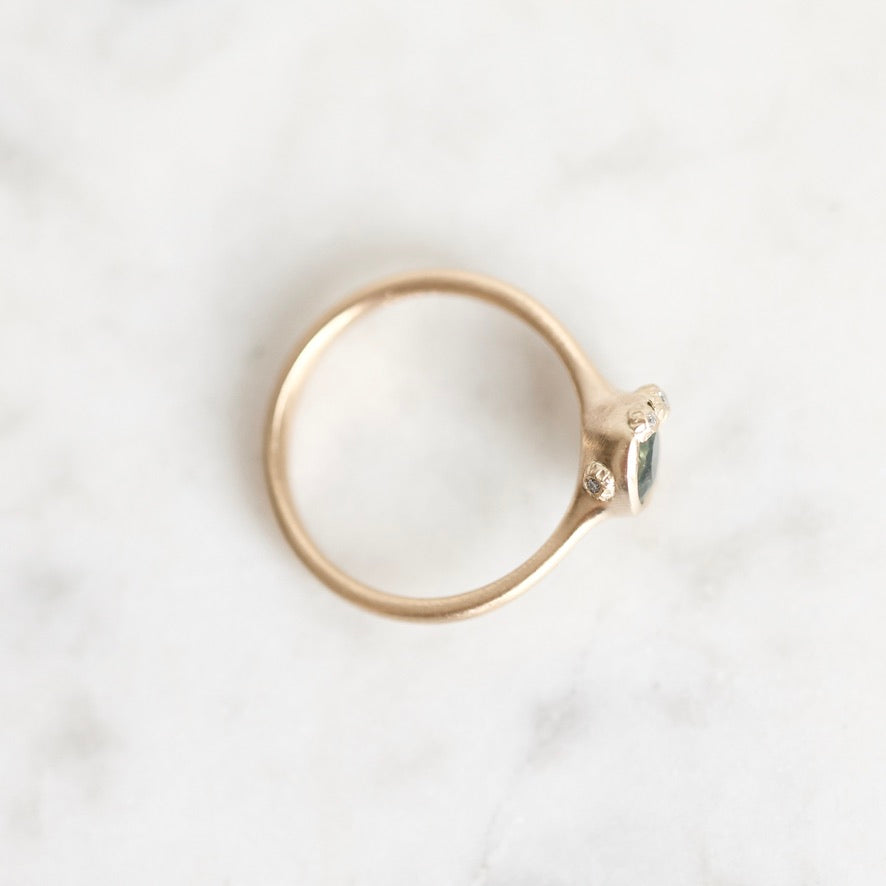 A delicate gold ring with a small marquise-cut green gemstone and a tiny clear accent stone, photographed from above on a white marble surface.