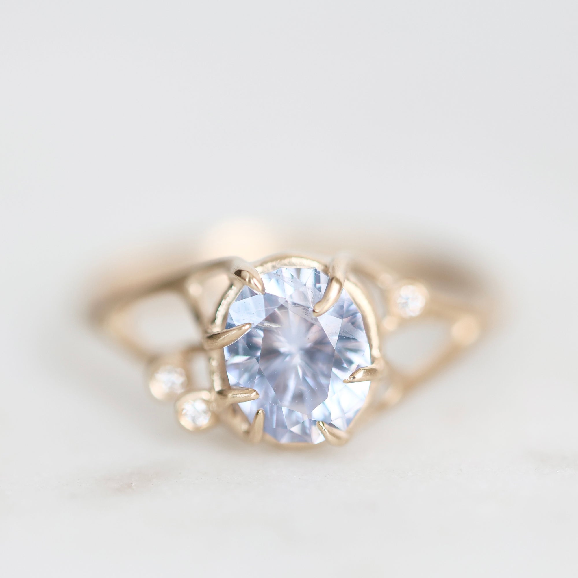 A delicate gold ring featuring a large oval-cut light blue gemstone in a prong setting, accented by two small round clear stones on the shoulders, photographed on a soft white background.