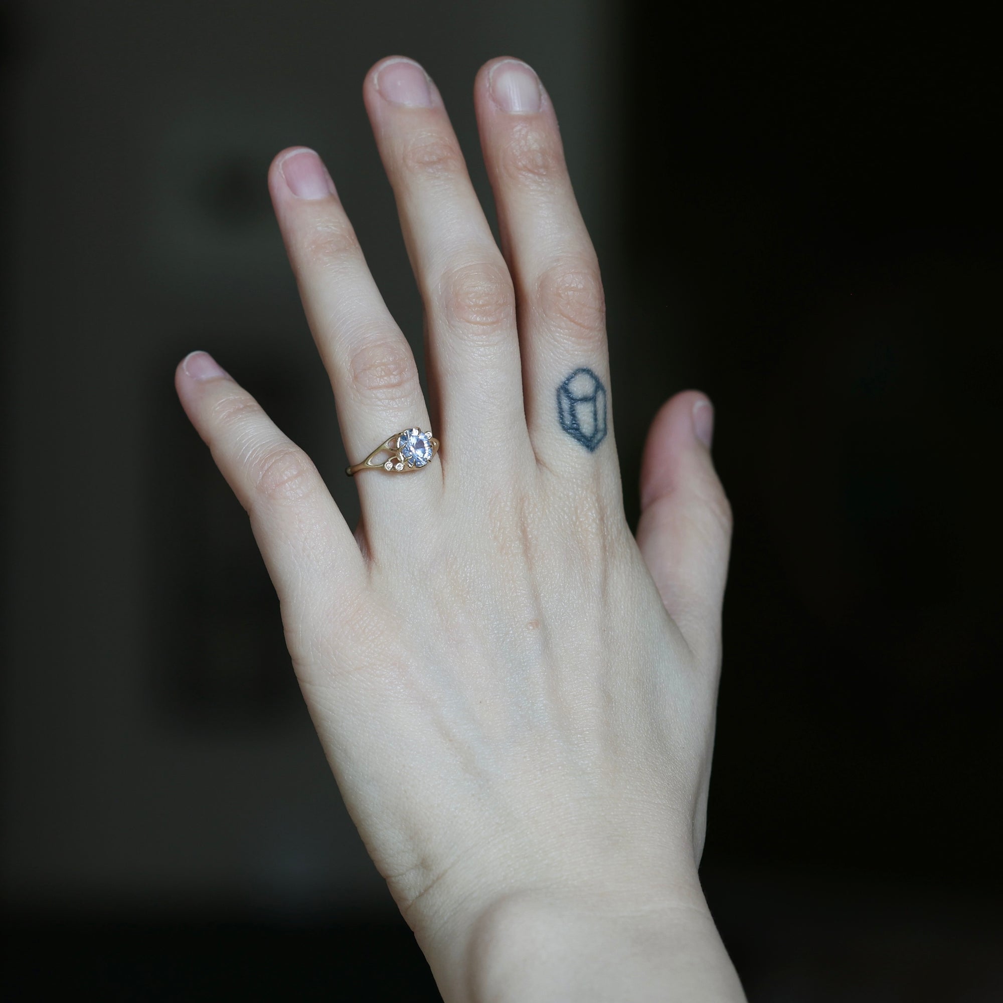A close-up of a person's hand wearing a delicate gold ring with a small clear gemstone, set against a dark, blurred background.