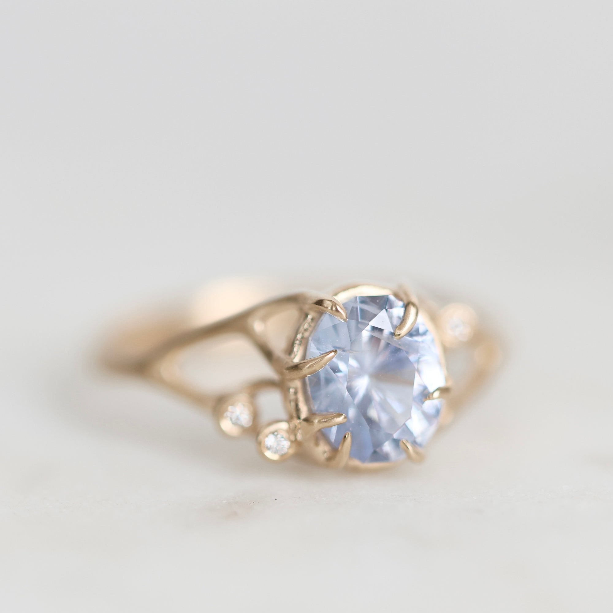 A delicate gold ring featuring a large oval light blue gemstone in a prong setting, accented with small round clear stones on the shoulders, photographed on a light gray background.