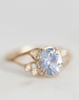 A delicate gold ring featuring a large oval light blue gemstone in a prong setting, accented with small round clear stones on the shoulders, photographed on a light gray background.