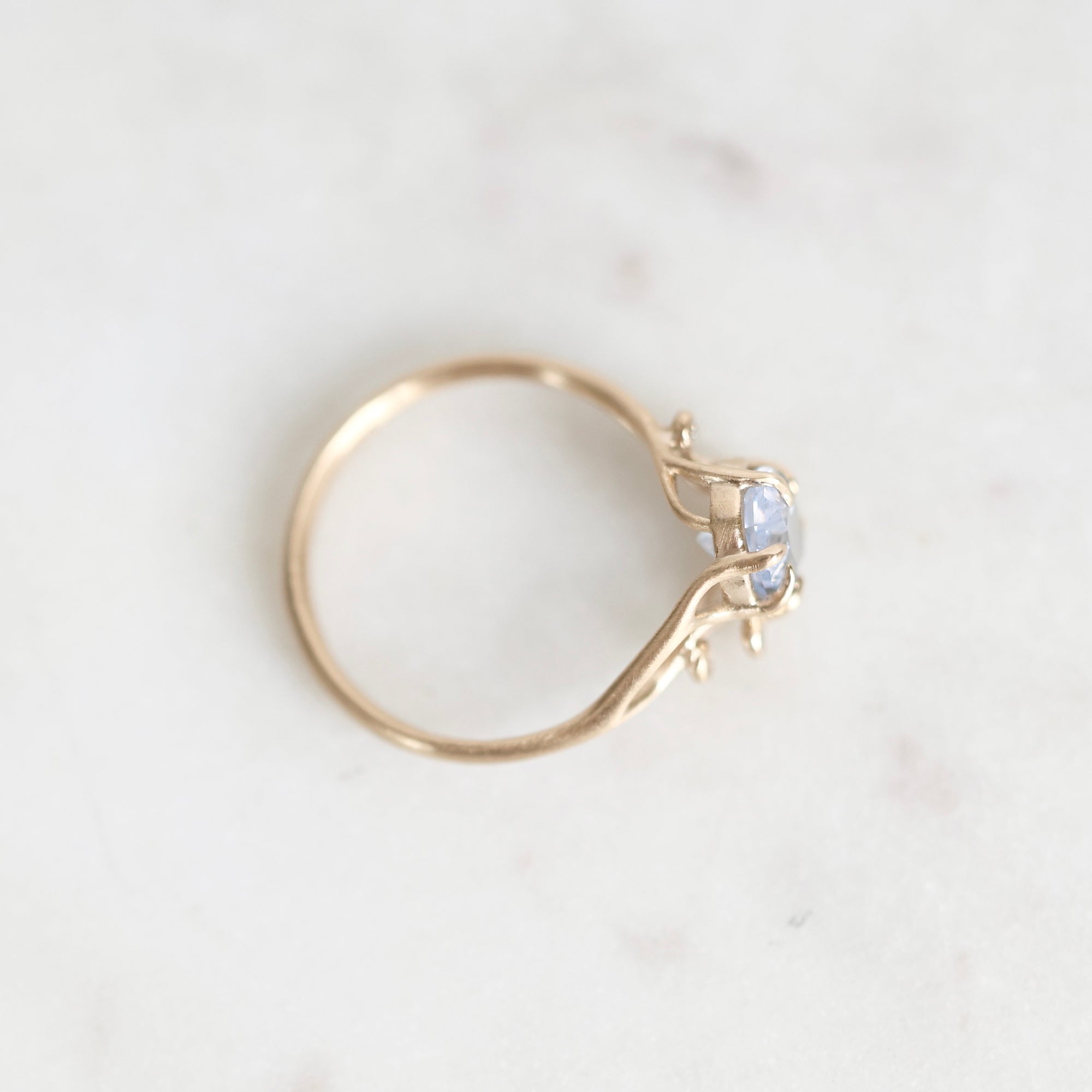 A delicate gold ring with a small light blue gemstone, photographed on a light marble surface.