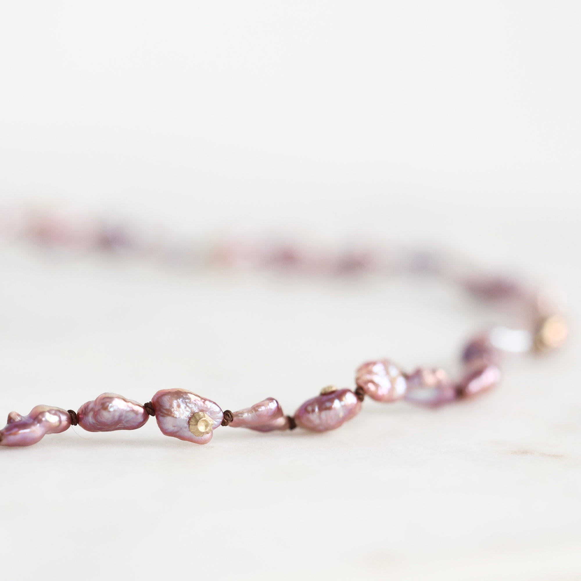A close-up of a beaded necklace made of irregular, lustrous baroque pearls in a dusty lavender-purple shade, strung on a dark cord against a bright, soft-focus white background.
