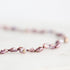 A close-up of a beaded necklace made of irregular, lustrous baroque pearls in a dusty lavender-purple shade, strung on a dark cord against a bright, soft-focus white background.