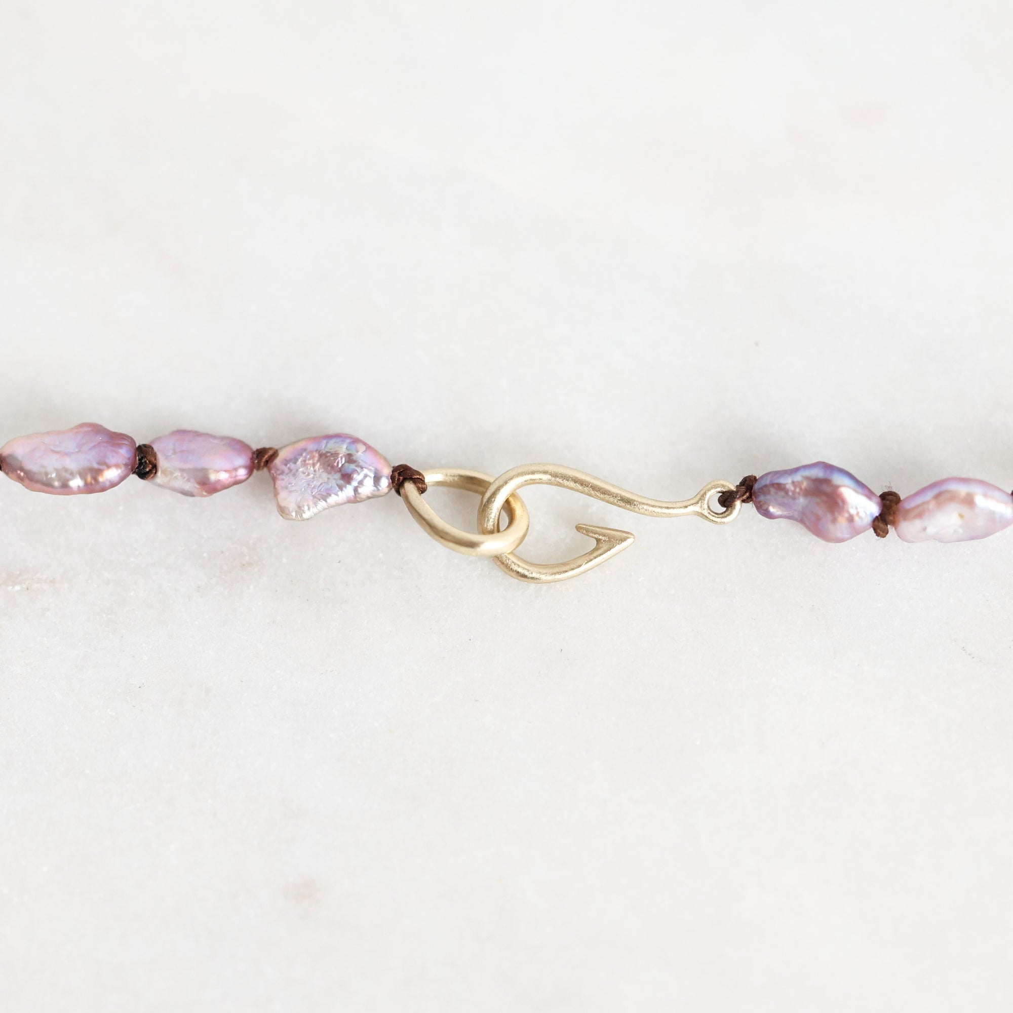 A close-up of a beaded necklace featuring irregular light purple baroque pearls threaded on a gold-toned chain with a decorative twist clasp, set against a plain off-white background.