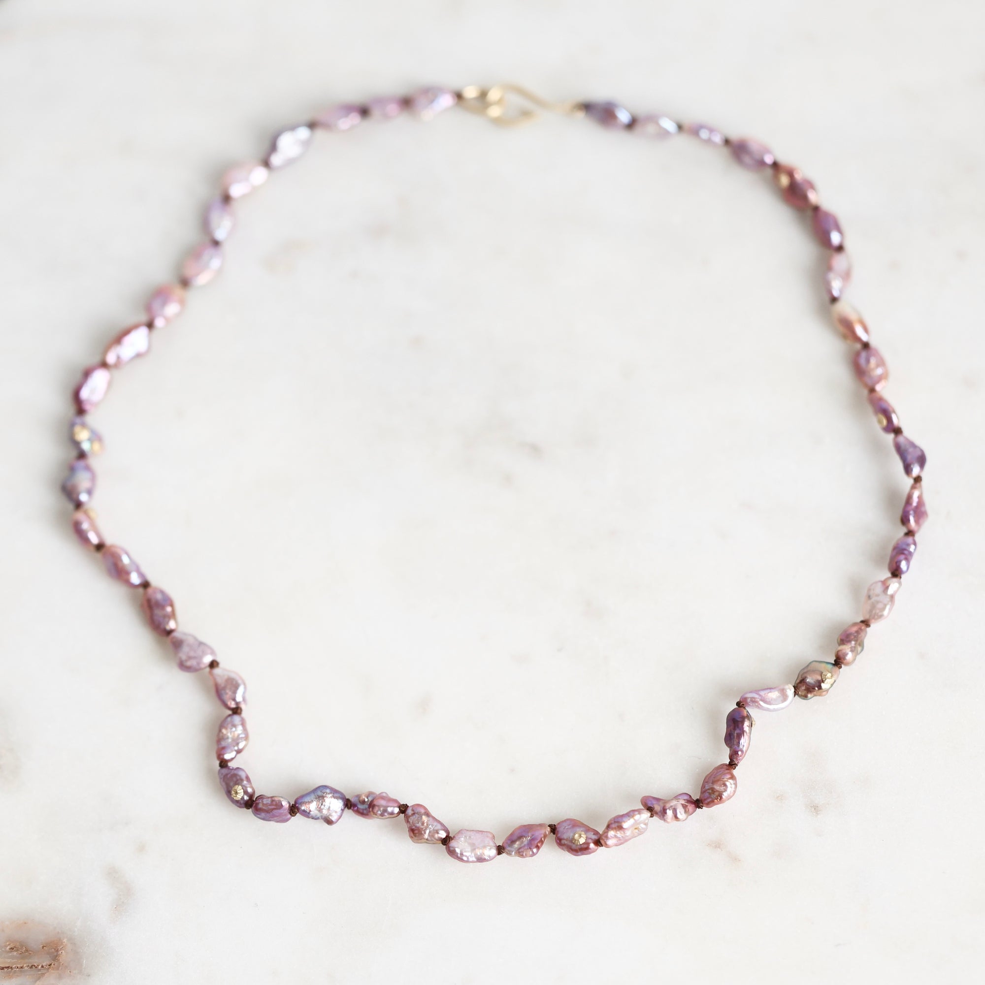 A baroque pearl necklace with irregular, faceted lavender-pink pearls and small gold spacer beads, displayed on a light marble surface.