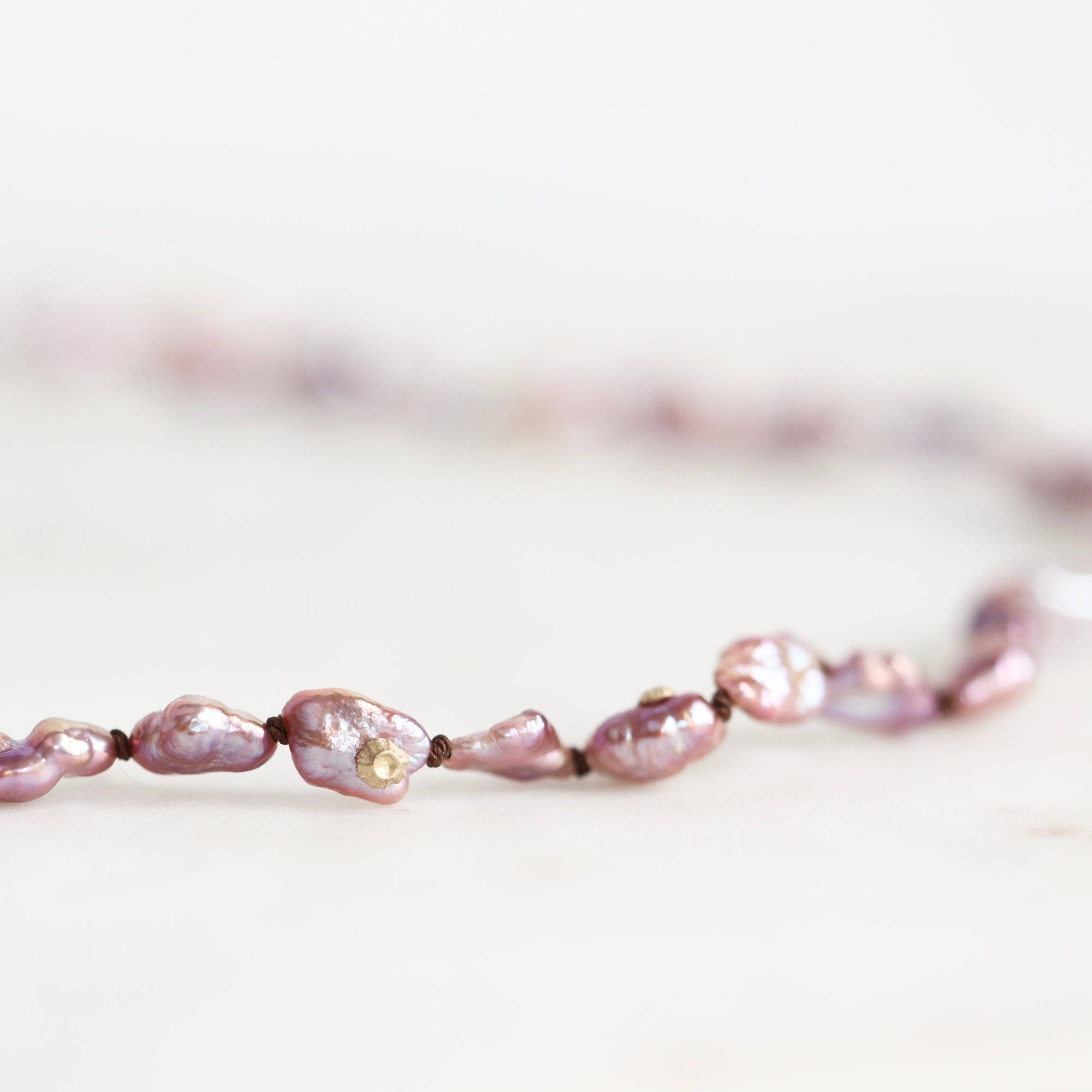 A close-up of a delicate necklace featuring irregular, baroque-style pinkish-purple freshwater pearls strung on a dark cord, resting on a light, neutral surface.