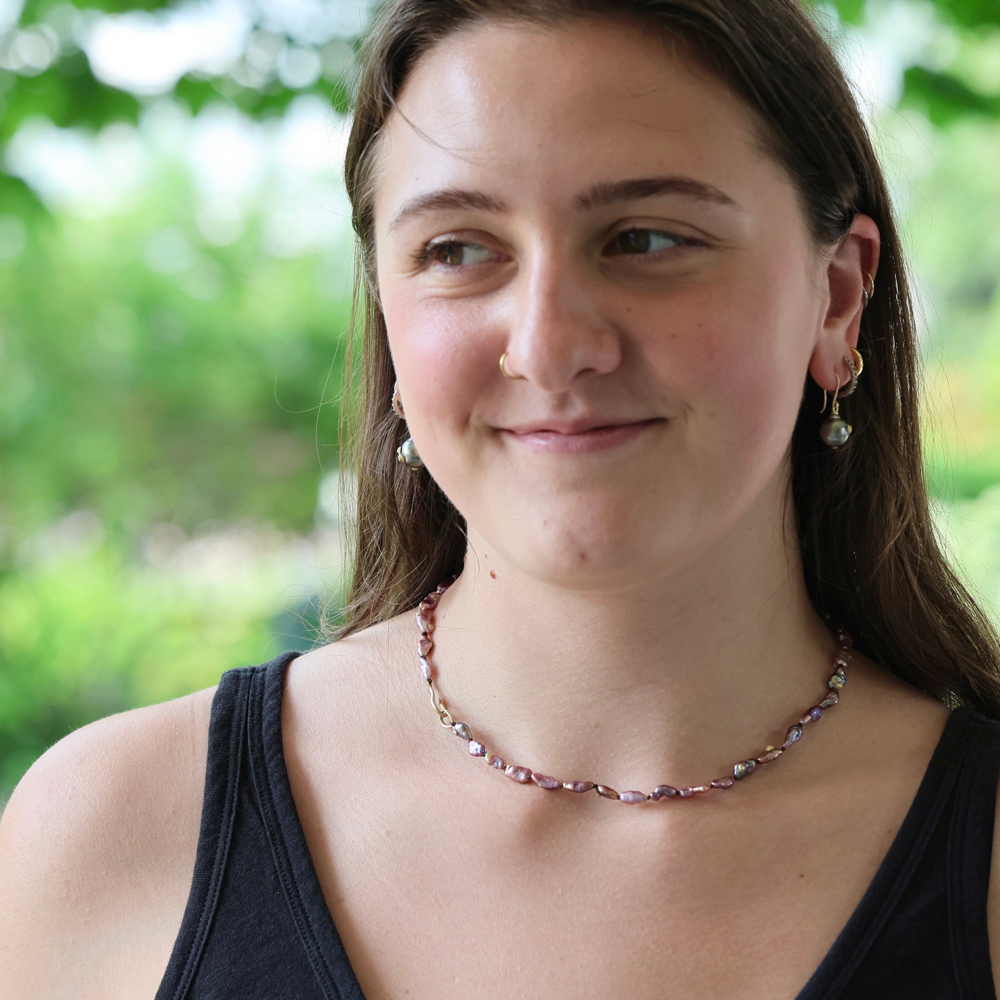 A woman with brown hair wearing a delicate beaded necklace with pink, purple, and white pearls, along with small gold hoop earrings, set against a soft-focus green outdoor background.