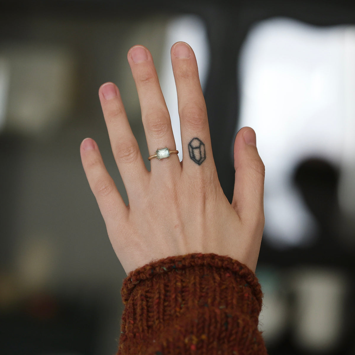 A close-up of a person's hand wearing two rings: a silver band with a small white oval stone on the ring finger, and a black geometric tattoo or stamp on the middle finger. The hand is wearing a brown knit sweater cuff, with a blurred dark background.