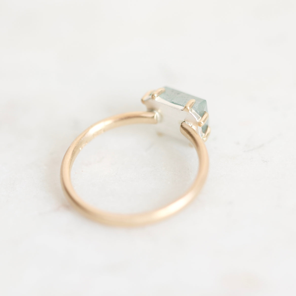 A minimalist open-band ring featuring a polished rose gold band and a small, faceted light green gemstone in a silver-toned prong setting, resting on a light marble surface.