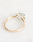 A minimalist open-band ring featuring a polished rose gold band and a small, faceted light green gemstone in a silver-toned prong setting, resting on a light marble surface.