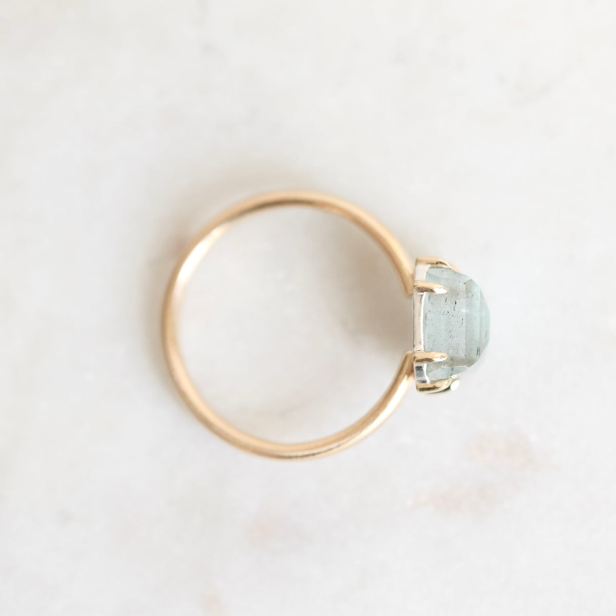 A minimalist gold ring featuring a small, bezel-set light blue stone, photographed from an overhead angle on a light, textured off-white surface.