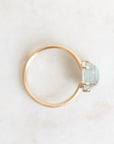 A minimalist gold ring featuring a small, bezel-set light blue stone, photographed from an overhead angle on a light, textured off-white surface.
