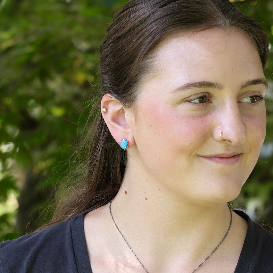 A close-up portrait of a woman outdoors wearing a small turquoise-blue stud earring, with green foliage blurred in the background.