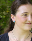A close-up portrait of a woman outdoors wearing a small turquoise-blue stud earring, with green foliage blurred in the background.