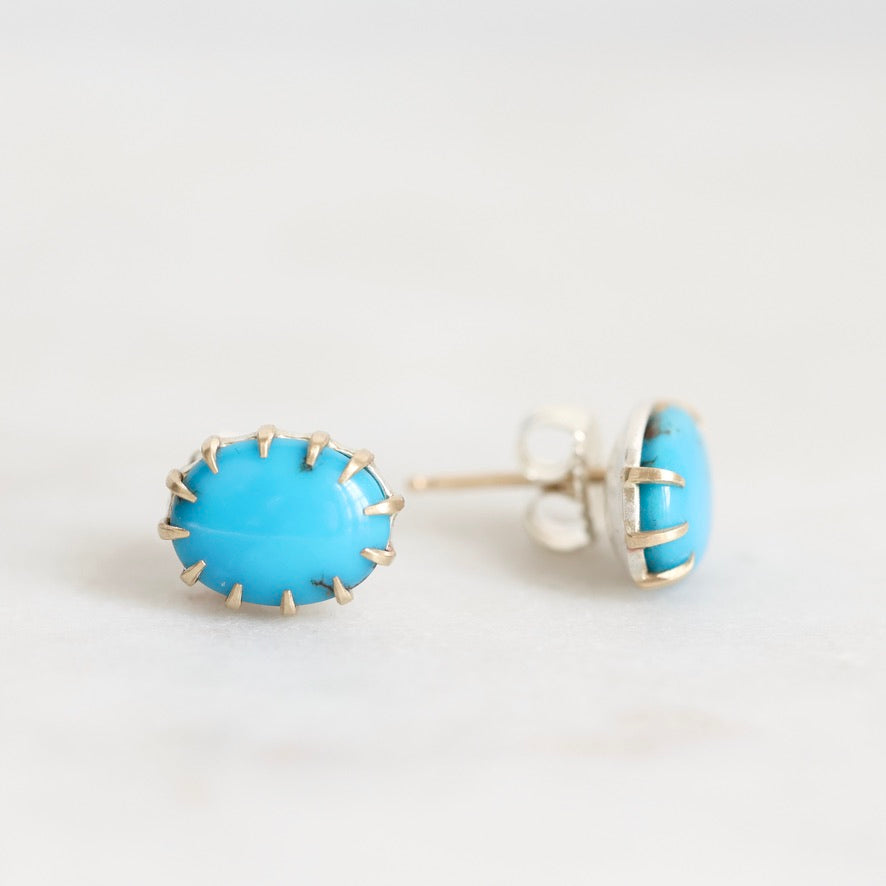 A pair of oval stud earrings featuring bright turquoise blue cabochon stones set in silver-toned metal with a textured, hand-hammered edge, shown on a light gray background.