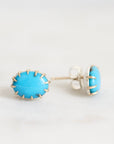 A pair of oval stud earrings featuring bright turquoise blue cabochon stones set in silver-toned metal with a textured, hand-hammered edge, shown on a light gray background.