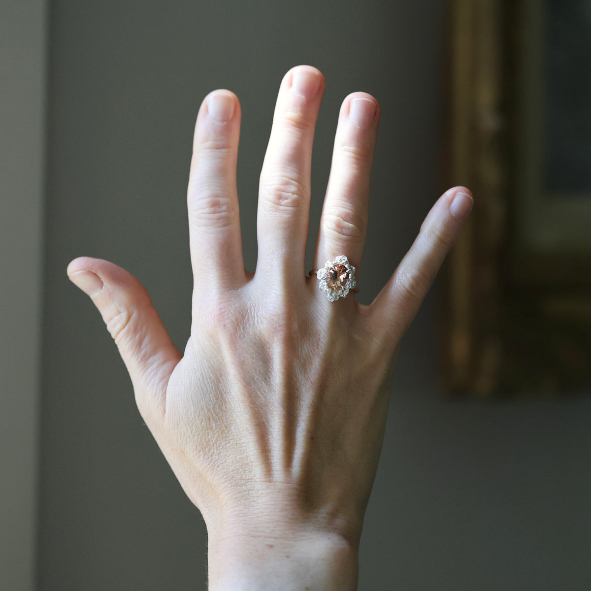 A close-up of a person's hand wearing a silver-toned ring with a floral cluster of clear gemstones, set against a softly blurred gray wall background.