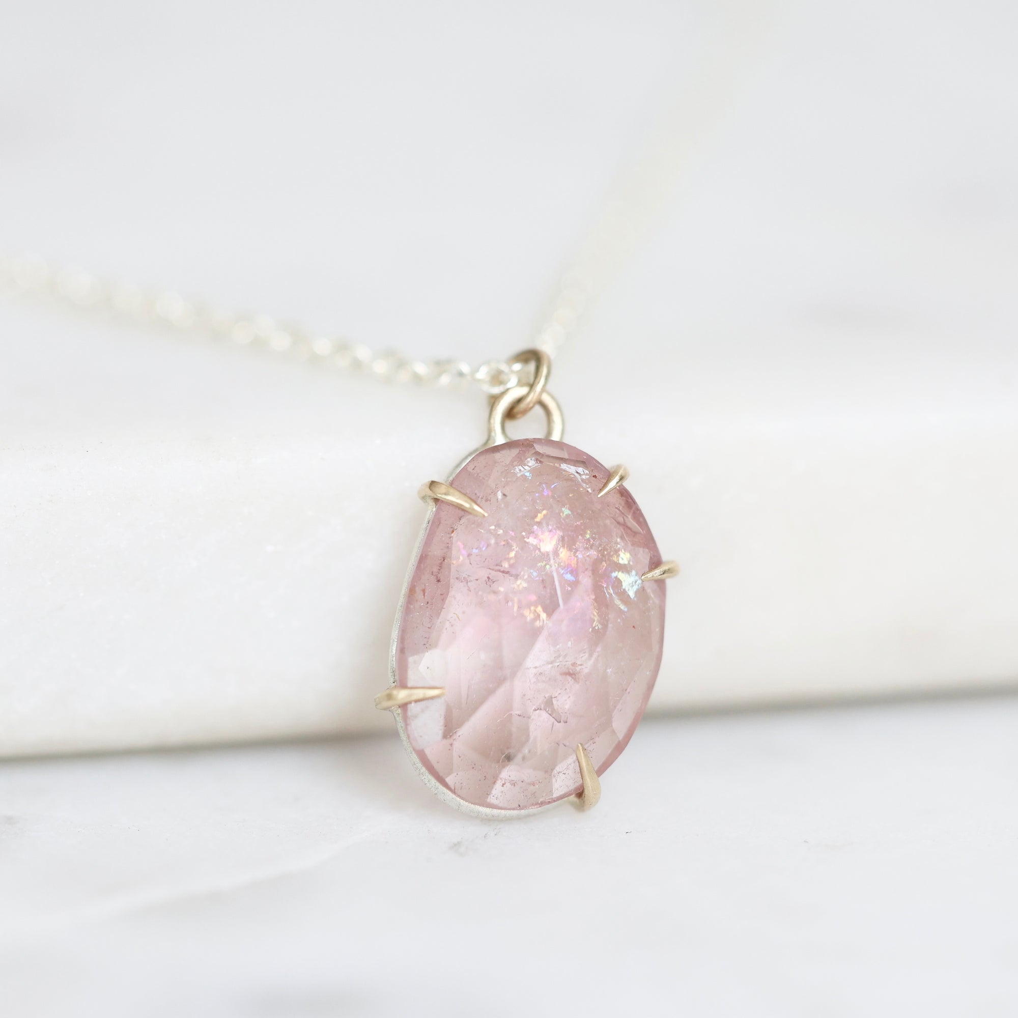 A close-up of a teardrop-shaped pink gemstone pendant in a gold-toned setting on a delicate chain, resting on a white marble surface.