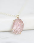 A close-up of a teardrop-shaped pink gemstone pendant in a gold-toned setting on a delicate chain, resting on a white marble surface.
