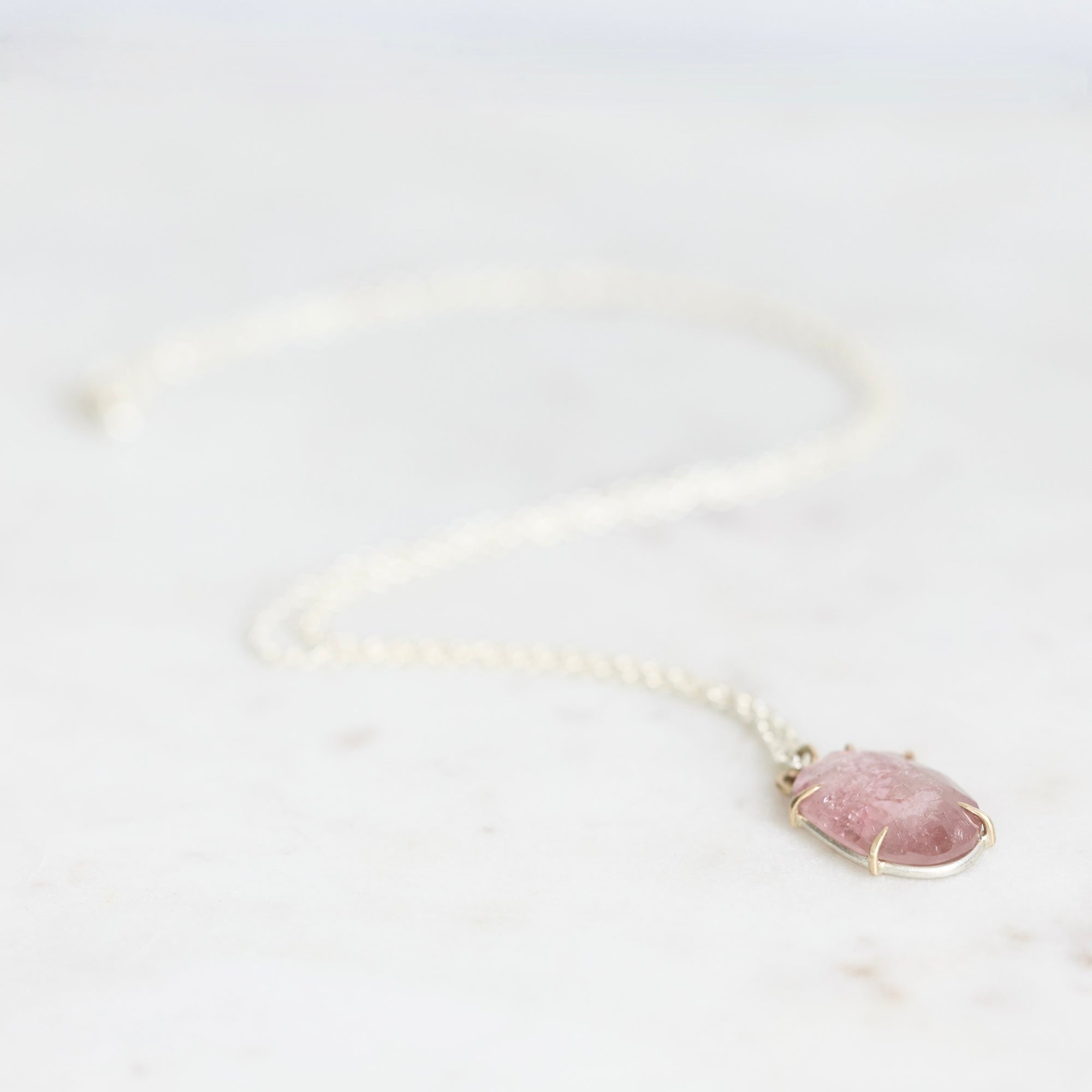 A delicate white beaded chain necklace with a small, oval-shaped pink gemstone pendant in a thin silver bezel setting, resting on a light marble surface.