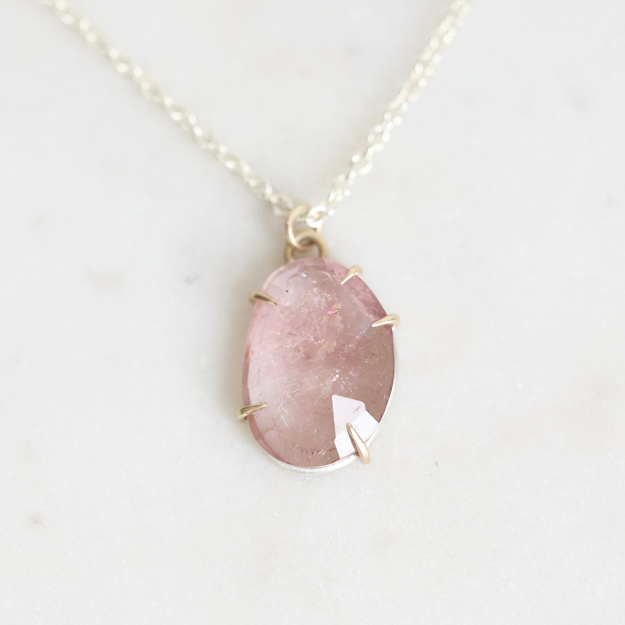 A delicate silver chain necklace featuring an irregular, teardrop-shaped pink gemstone pendant in a simple silver bezel setting, displayed on a light marble surface.