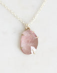 A delicate silver chain necklace featuring an irregular, teardrop-shaped pink gemstone pendant in a simple silver bezel setting, displayed on a light marble surface.