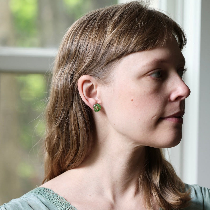 A woman in profile wearing a small green flower-shaped stud earring, photographed indoors near a window.