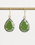 A pair of gold drop earrings with faceted green teardrop gemstones, hanging from a gold rod against a light gray background.