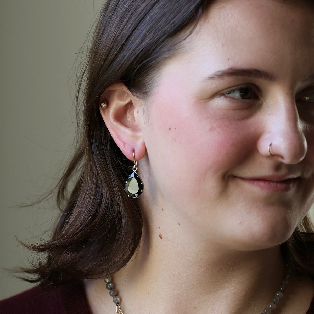 A close-up of a woman wearing a teardrop-shaped dangle earring with a pale green stone and dark blue accents, shown against a plain beige background.