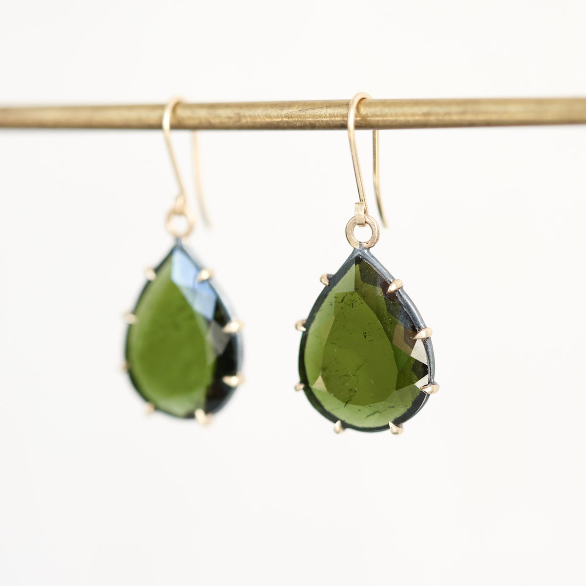 Teardrop-shaped green gemstone drop earrings in gold settings hanging from a gold display rod against a white background.