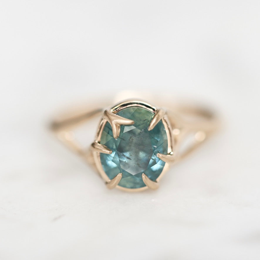 A gold ring with a round teal-blue gemstone in a prong setting, photographed close-up on a light marble-like surface.