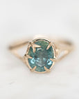 A gold ring with a round teal-blue gemstone in a prong setting, photographed close-up on a light marble-like surface.